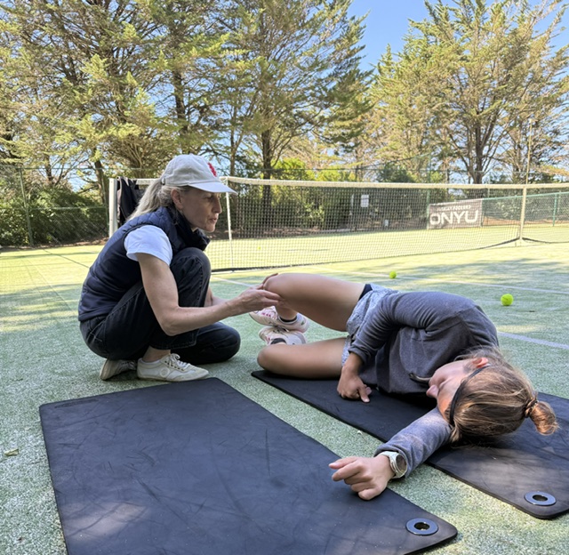 Naomi guiding a young athlete's leg movement on a mat beside a tennis court