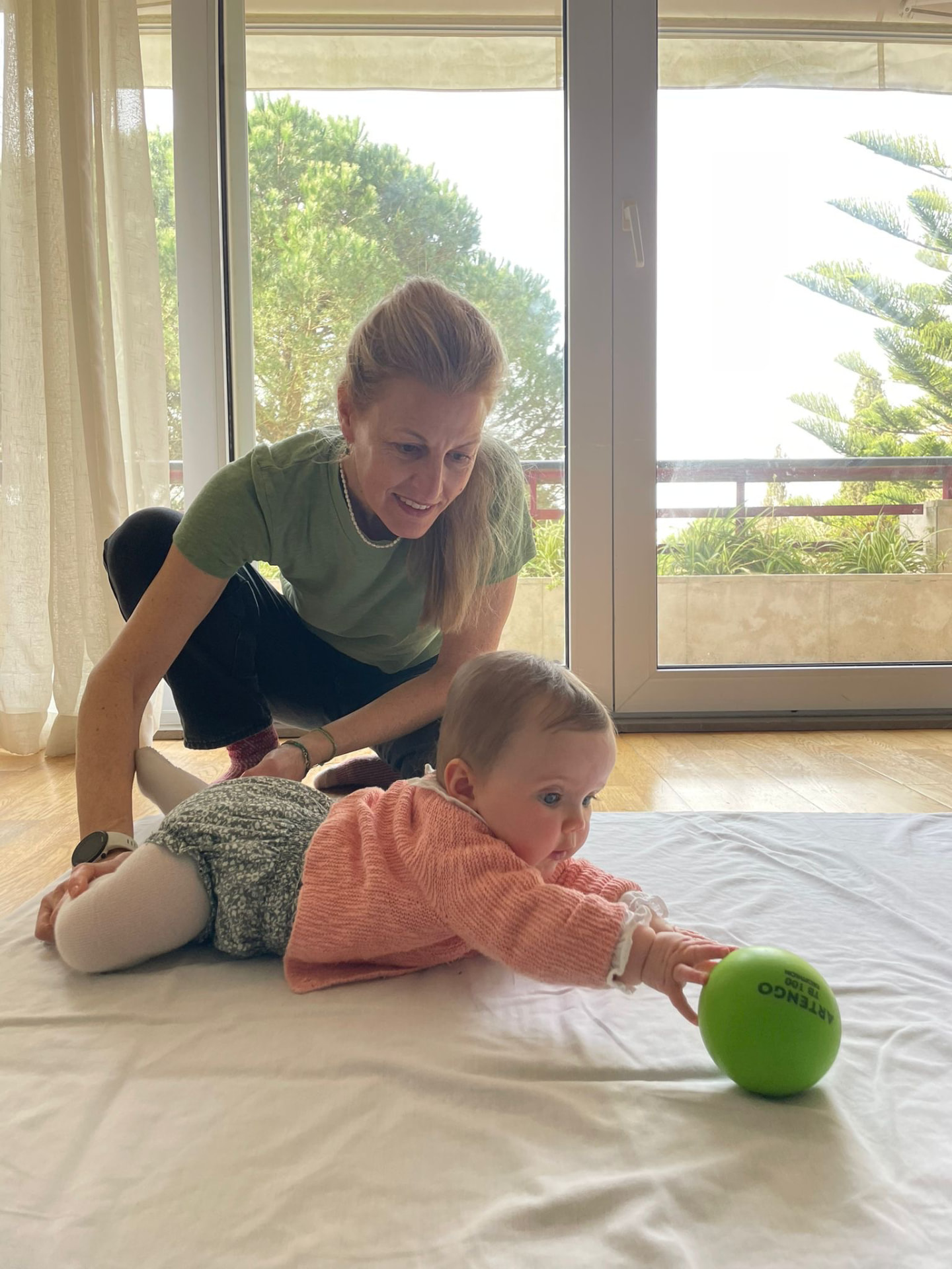 Naomi beside a baby on the floor as the baby reaches forward for a green ball